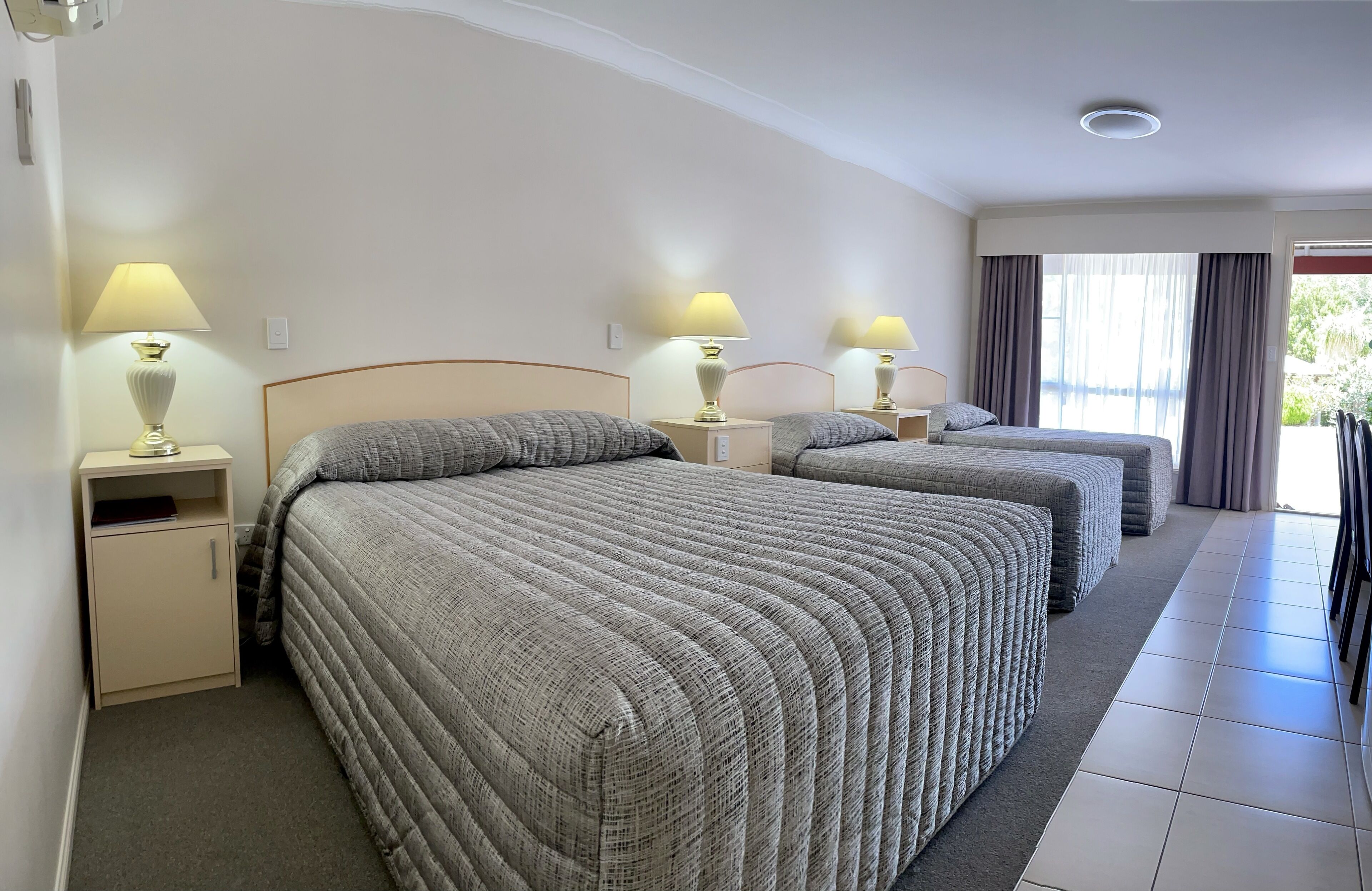 A hotel room with two beds, each with a bedside table and lamp, a small cabinet, and a view of the outdoors through a window with curtains.