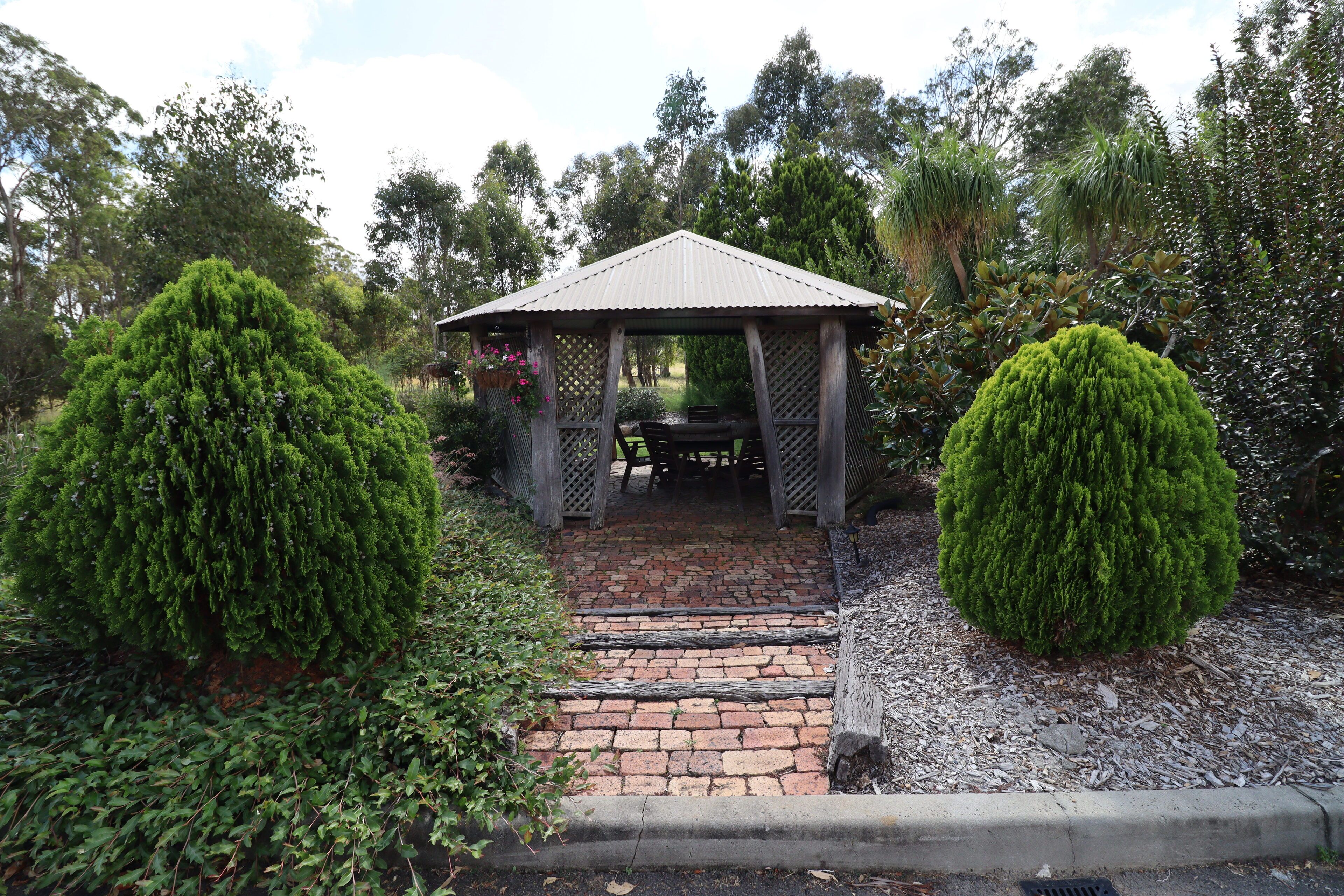 A gazebo with a tiled roof, surrounded by lush greenery and a brick pathway.