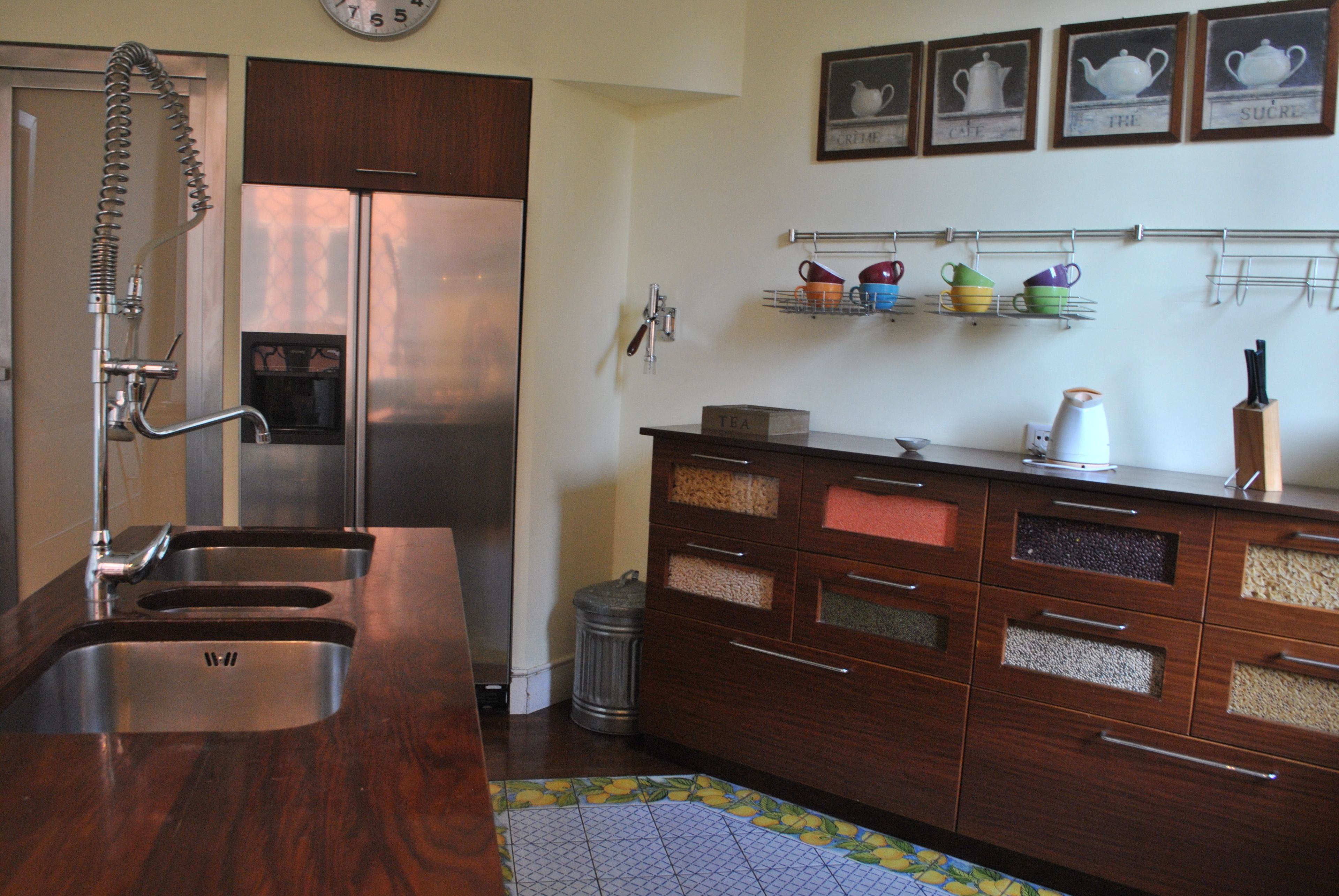 A kitchen with a stainless steel refrigerator, double sinks, wooden cabinets with multiple drawers, and a small trash can.