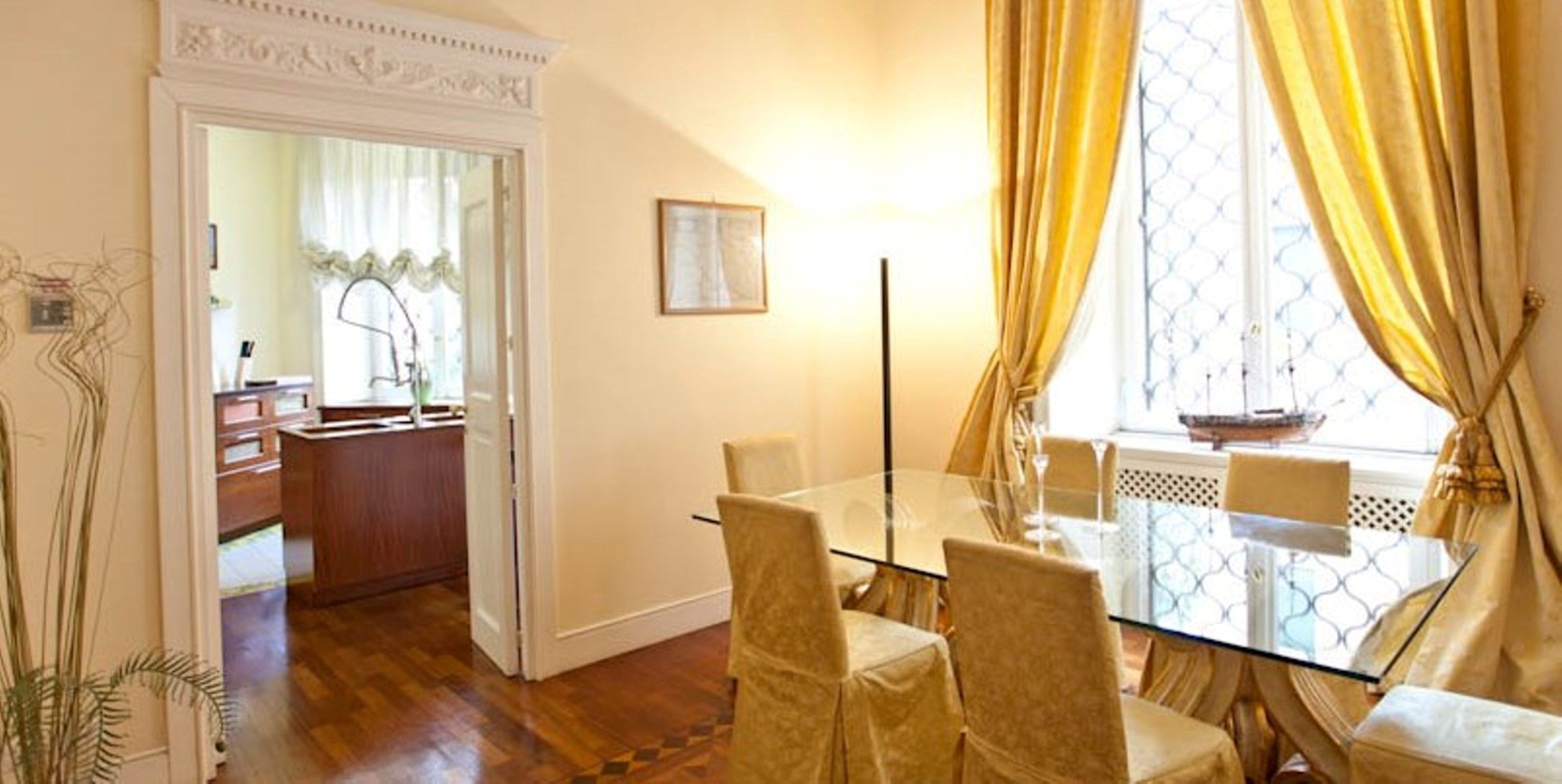 Un coin repas avec une table et des chaises en verre, une cuisine visible à travers une porte ouverte et une fenêtre avec des rideaux.