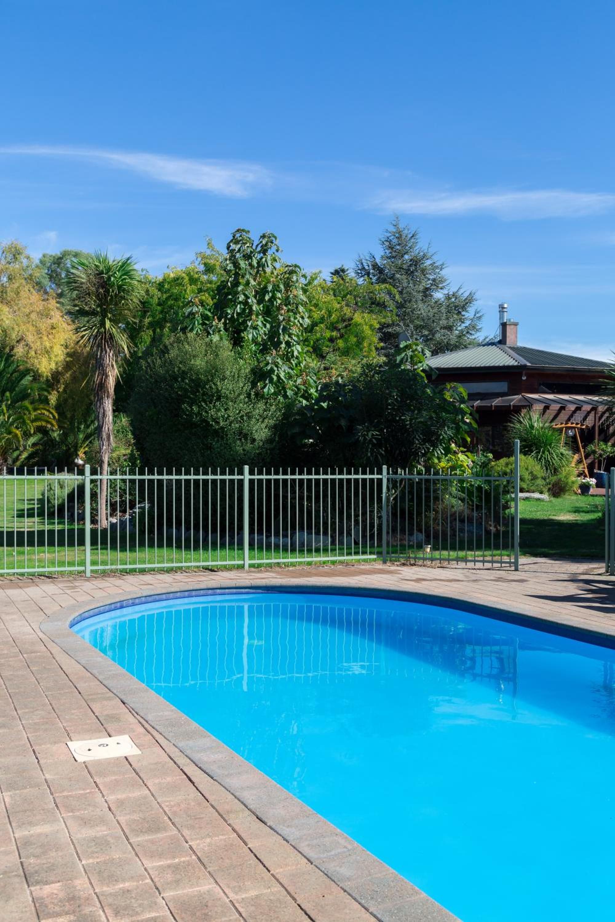 Piscina com revestimento de tijolos, cercada por grade de metal, árvores e uma casa com telhado verde.