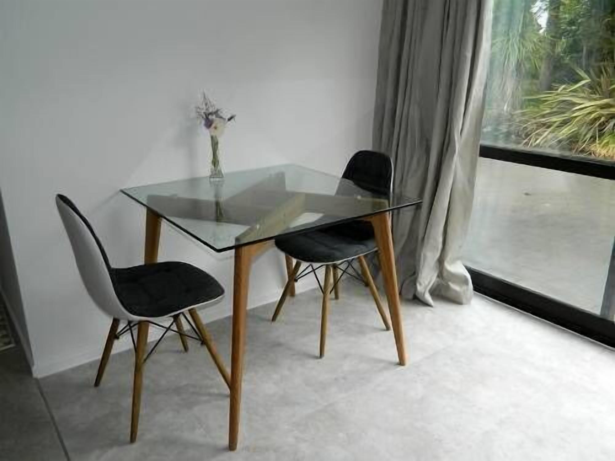 Et moderne spiseområde med et glasbord og træstole, en vase med blomster og udsigt til grøn vegetation gennem et vindue.