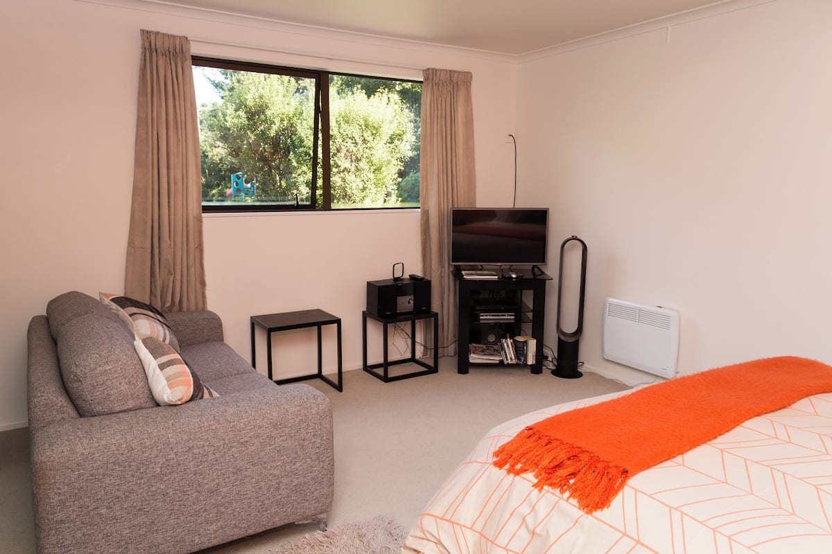 A bedroom with a sofa, a bed with an orange blanket, a TV, a small table, and a radiator.