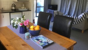 A kitchen with white cabinets, a microwave, and a kettle. There is a dining table with a vase of flowers and a bowl of lemons. A television is visible in the background.