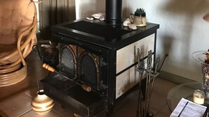 A wood-burning stove with a black chimney, a woven basket, and a potted plant in a room with a wooden floor and a large plant in the corner. - The Blanco River is calling your name!! Come and enjoy!! (Wimberley)