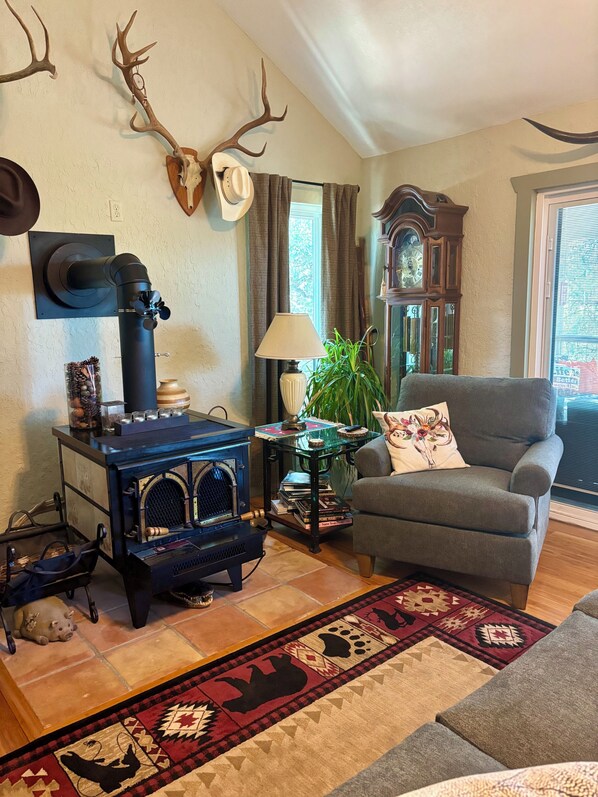 A cozy living room with a vintage stove, a comfortable armchair, a decorative wall with antlers, a clock, and a large window.