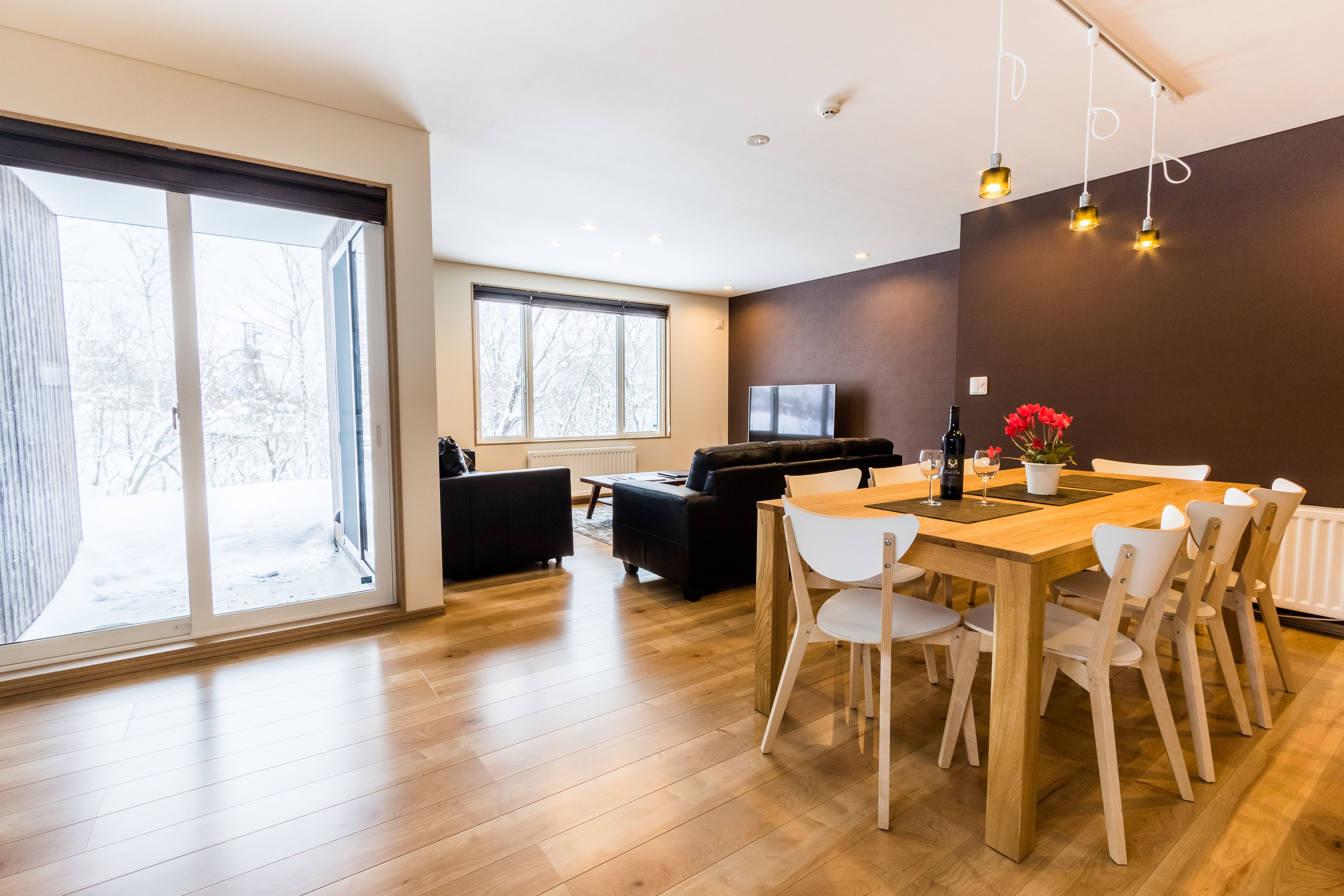 A modern living room with a large window, a sofa, a TV, and a dining area with a wooden table and chairs.
