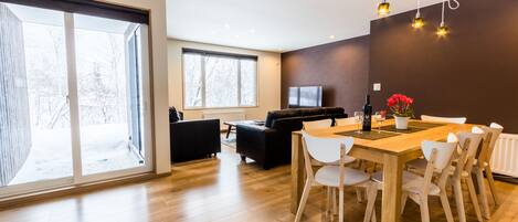 A modern living room with a large window, a sofa, a TV, and a dining area with a wooden table and chairs.