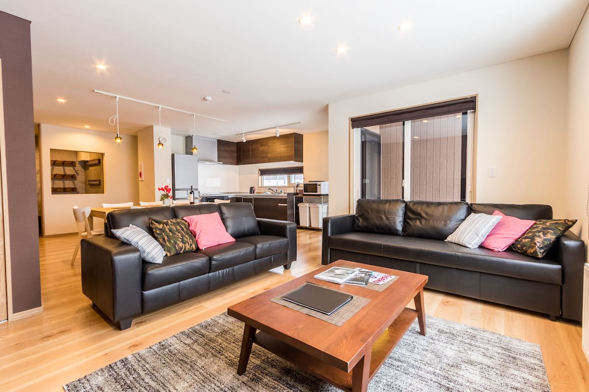A modern living room with two black leather sofas, a wooden coffee table, and a kitchen in the background.