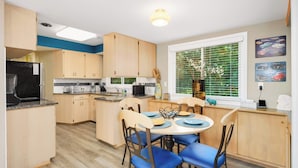 Une cuisine de taille réduite, dotée de meubles en bois, d’une table à manger avec des chaises bleues et d’une fenêtre avec des stores.