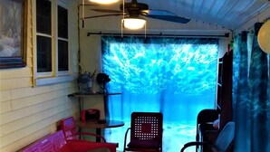 Un porche cubierto con un sofá rojo, una silla negra, un enfriador de Coca-Cola y un cubo azul con herramientas de jardinería.