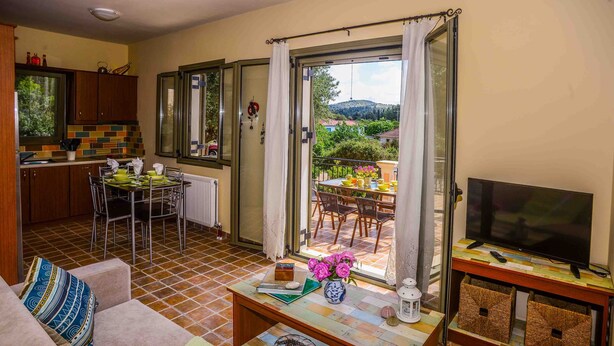 A living room with a sofa, a coffee table, and a TV. There is a dining area with a table and chairs. The room has a view of a garden and hills. - #1-Clean, self catering 2 bedroom apartment with large balcony and free parking (Antipata)