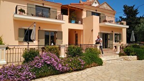 A two-story building with balconies, a stone-paved entrance, and purple flowering plants. - #1-Clean, self catering 2 bedroom apartment with large balcony and free parking (Antipata)