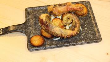 Une assiette contenant un gigot d’agneau rôti, des pommes de terre et un petit objet rond, le tout présenté sur une assiette grise à texture.