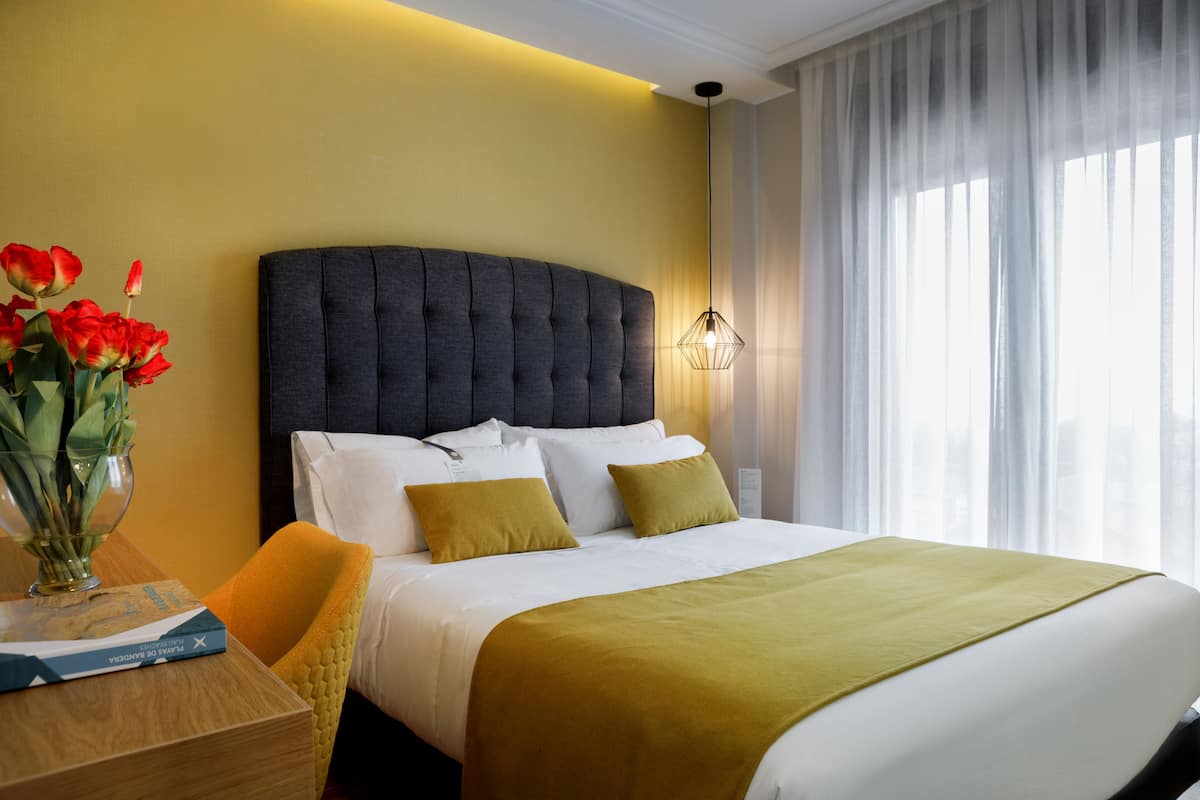 A hotel room with a large bed, a bedside table with a book, a vase of flowers, and a window with curtains.
