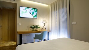A hotel room with a bed, a desk with a chair, a TV mounted on the wall, and a window with curtains.