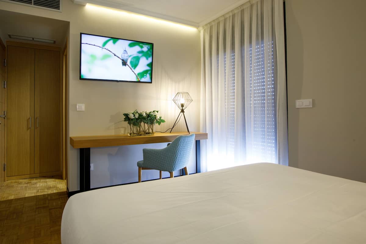 A hotel room with a bed, a desk with a chair, a TV mounted on the wall, and a window with curtains.