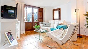 Een woonkamer met een witte bank, een glazen salontafel en een witte fauteuil. Er hangt een televisie aan de muur en op de tafel staat een plant.