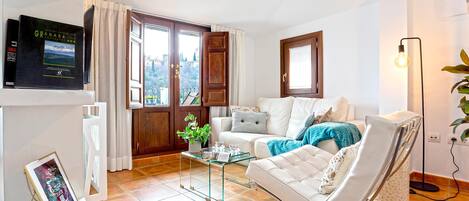 A living room with a white sofa, a glass coffee table, and a white armchair. There is a TV mounted on the wall and a plant on the table.