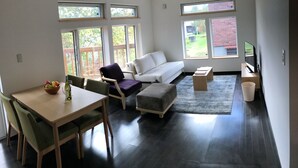 A modern living room with a dining area, large windows, a white sofa, and a flat-screen TV. - Lodge Mori Apartments (Kutchan)