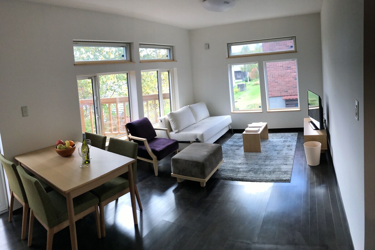 A modern living room with a dining area, large windows, a white sofa, and a flat-screen TV.