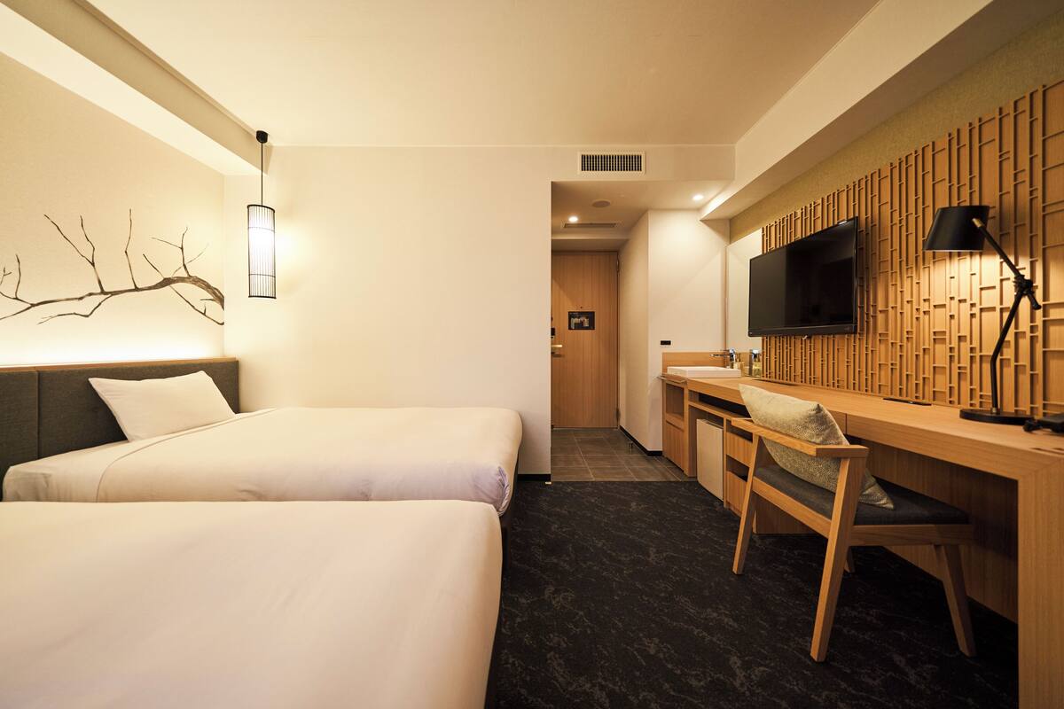 A hotel room with two beds, a desk, a chair, a TV, and a wall-mounted light fixture.