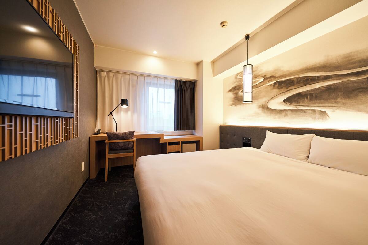 A modern hotel room with a large bed, a desk with a chair, a flat-screen TV, and a large abstract painting on the wall.