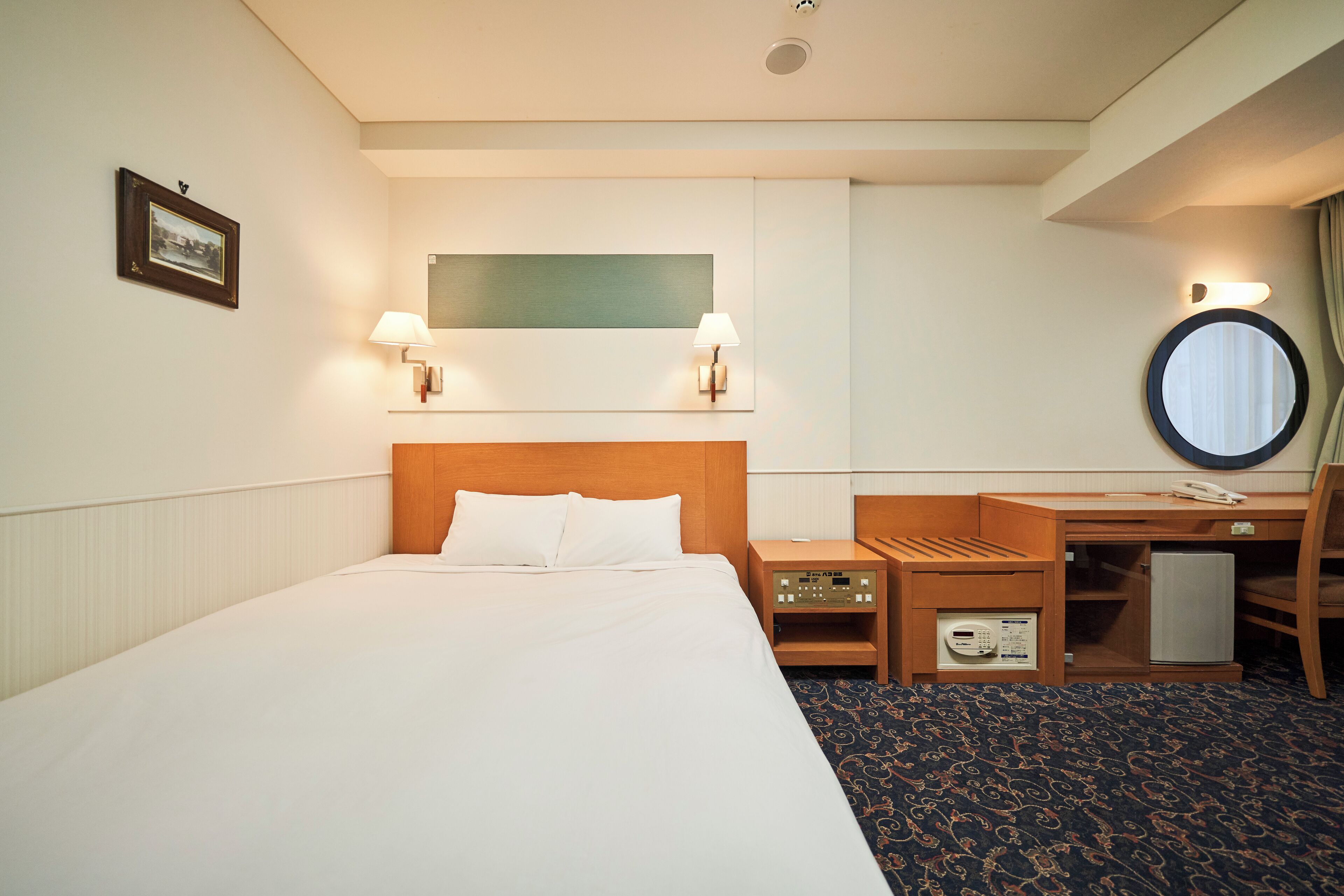 Habitación de hotel con una cama grande, mesitas de noche con lámparas, un escritorio, una silla, un refrigerador pequeño y un espejo adosado a la pared.