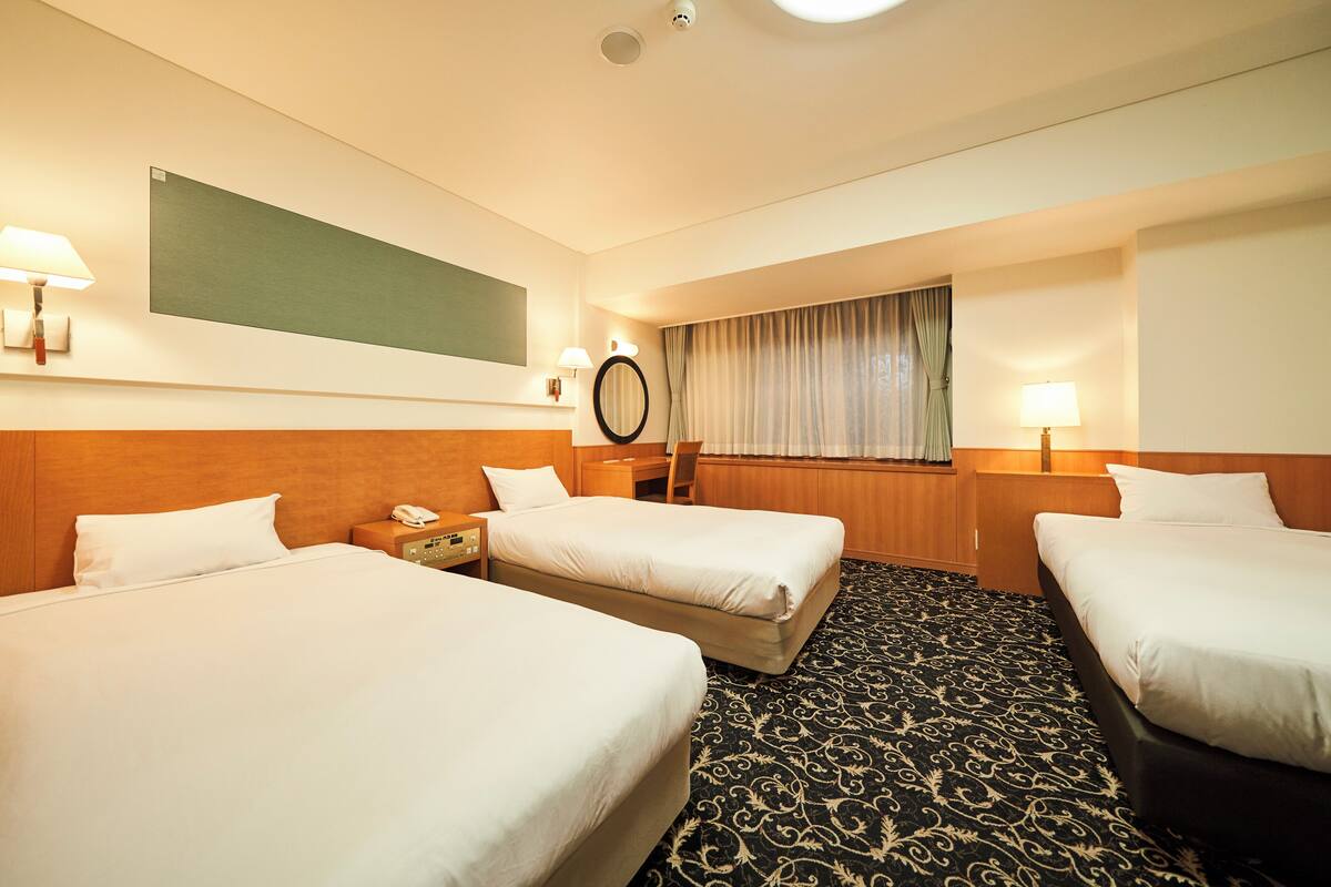 A hotel room with three beds, a desk, a chair, a telephone, a mirror, and a window with curtains.