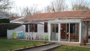 Una casa con techo de tejas, una puerta de cristal que da a un patio con sillas de plástico y una mesa, y un gato acostado sobre el césped.
