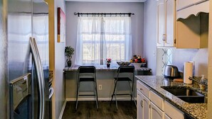A kitchen with a refrigerator, two bar stools, a window with sheer curtains, and wooden flooring.