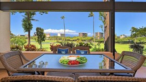 A glass-top table with a salad and a bottle of water, surrounded by wicker chairs, on a balcony with a view of a golf course and buildings. - Amazing Views; Walk to Kaanapali Beach and Whaler Village - Kaanapali Royal N102 (Lahaina)
