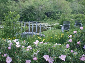 A garden with pink flowers, a wooden bridge, and two Adirondack chairs.