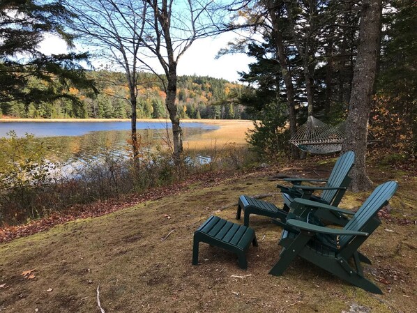 A lakeside setting with Adirondack chairs and a hammock, surrounded by trees and a view of the lake.