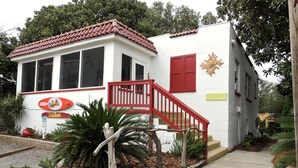 Un edificio blanco con techo rojo y una escalera roja que lleva a una puerta. Hay un letrero con un sol y una palmera en la fachada.