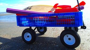 Un carro azul con ruedas en la playa, que contiene una tabla de surf, una pala y un objeto rojo.