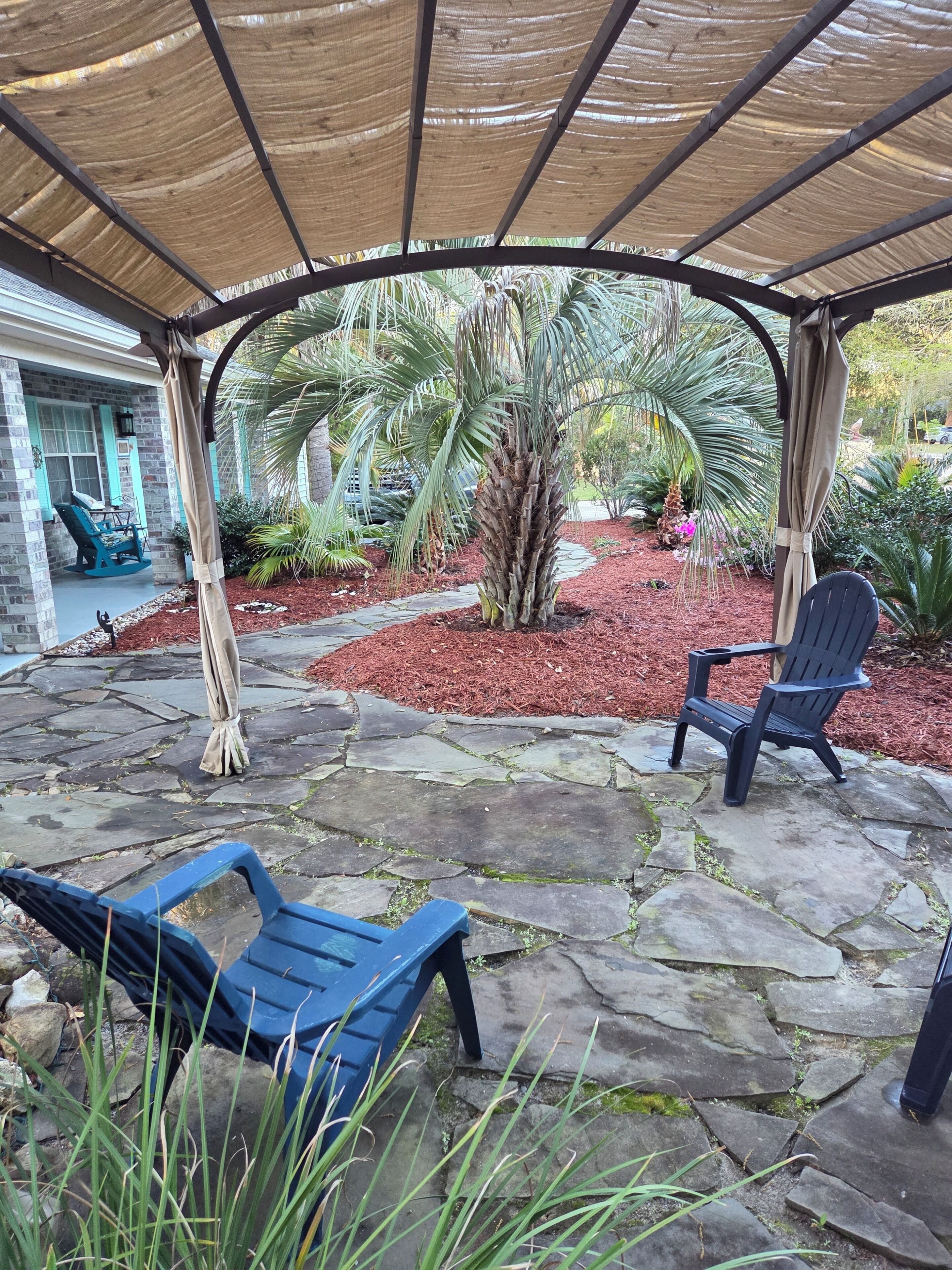 A covered patio with stone flooring, two blue chairs, and a palm tree.