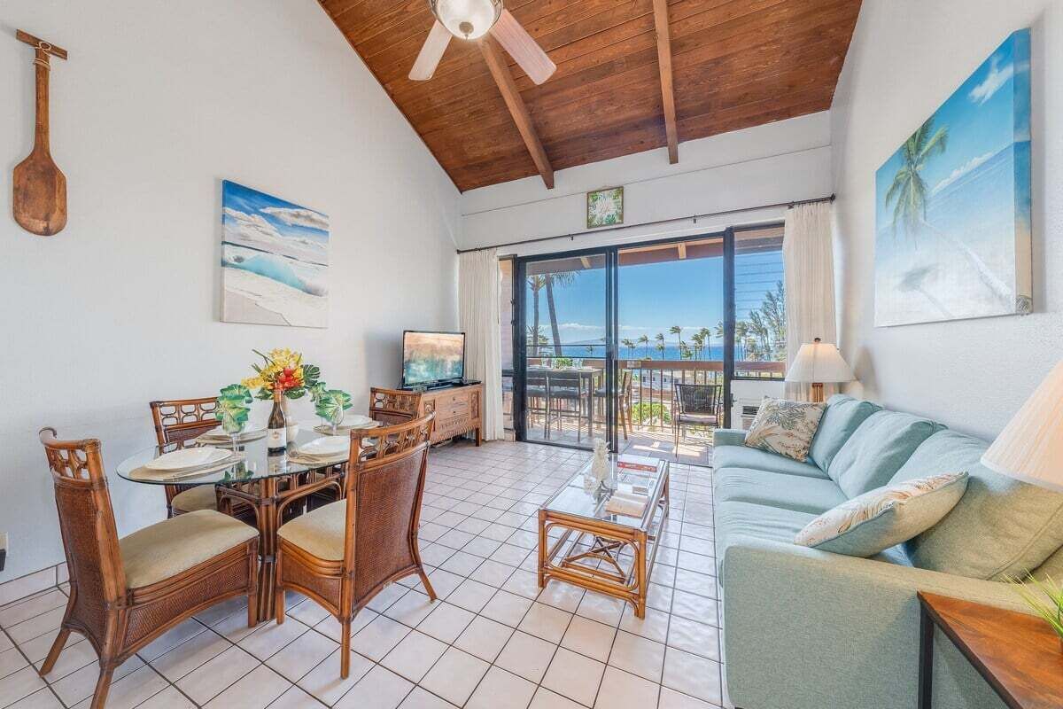 A spacious living area with a dining table, chairs, and a sofa. There is a ceiling fan, a television, and a view of the ocean through large windows.