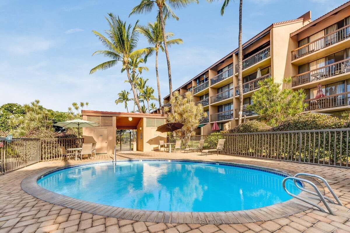 A swimming pool with lounge chairs and umbrellas, surrounded by palm trees and a multi-story building with balconies.