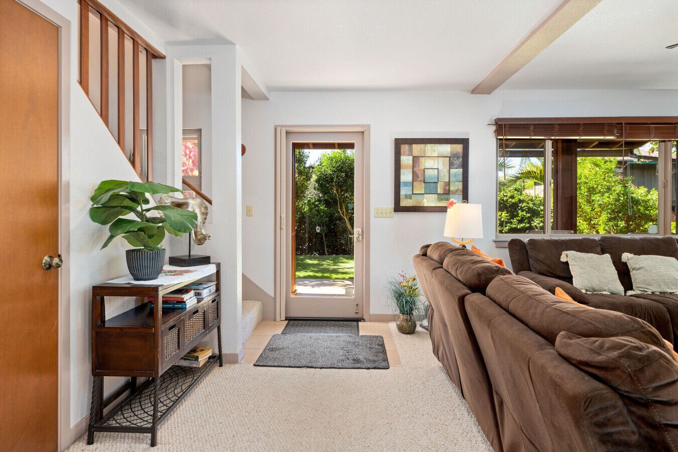 A living room with a brown sofa, a wooden staircase, a door leading to a garden, and a small table with a plant.