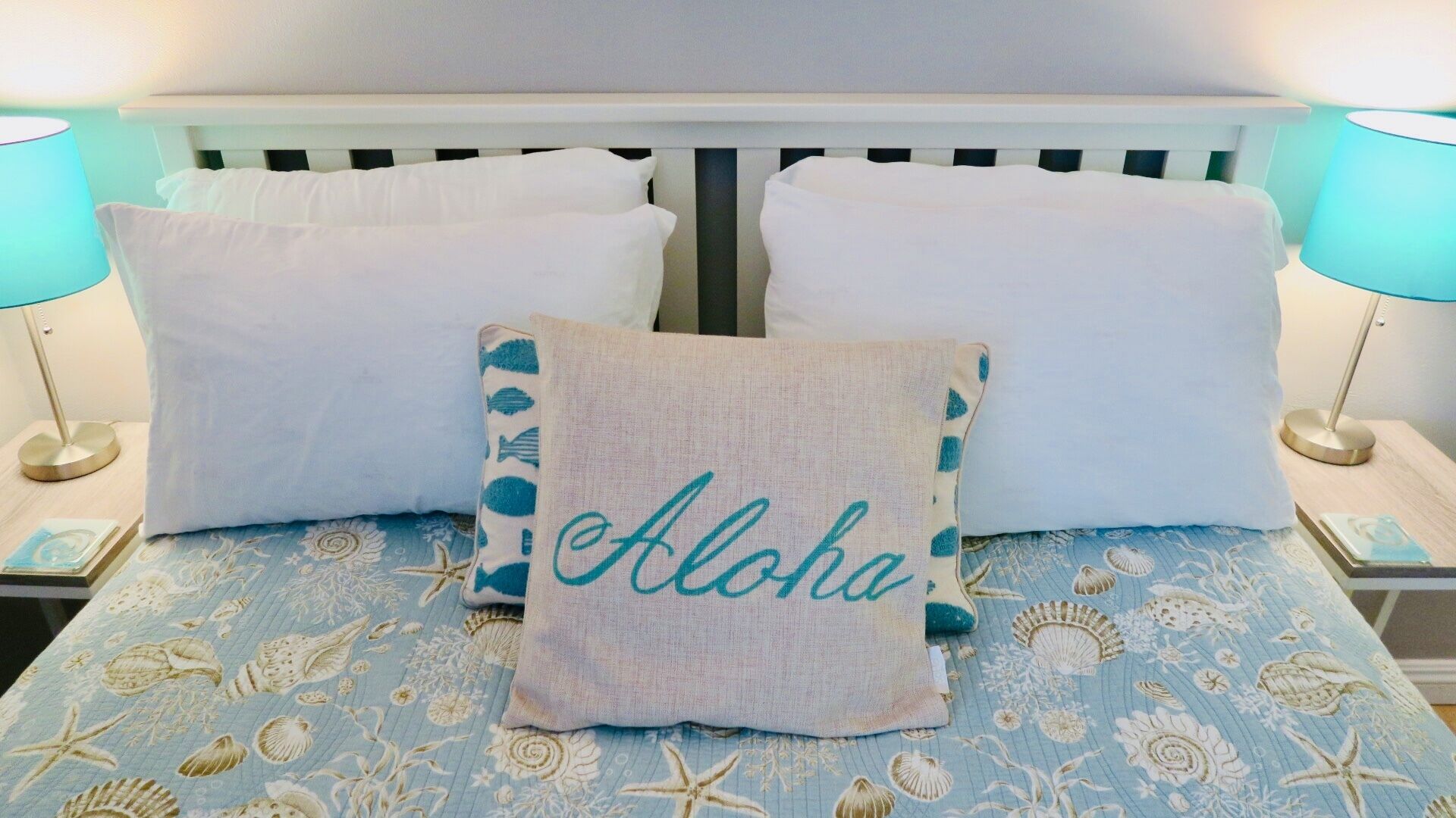 Una cama con una colcha azul estampada con flores, almohadas blancas y una almohada decorativa con la palabra 'Aloha'.