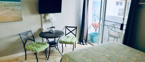 A hotel room with a bed, two chairs, a small table, a TV, and a balcony with a view of buildings.