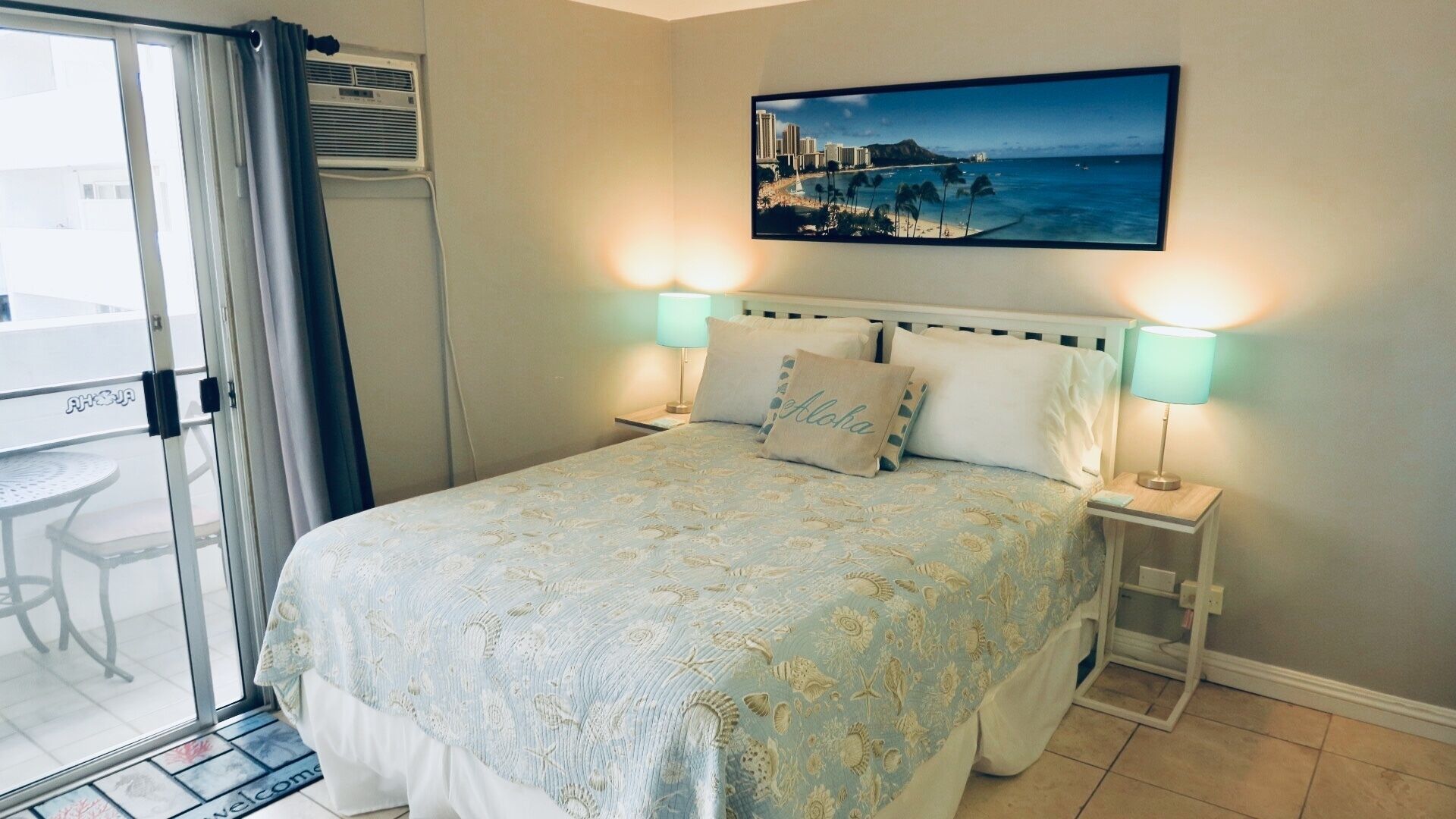 Ein Schlafzimmer mit einem Bett, zwei Nachttischlampen, einem Fenster mit Vorhängen und einem Bild von einer Strandlandschaft.