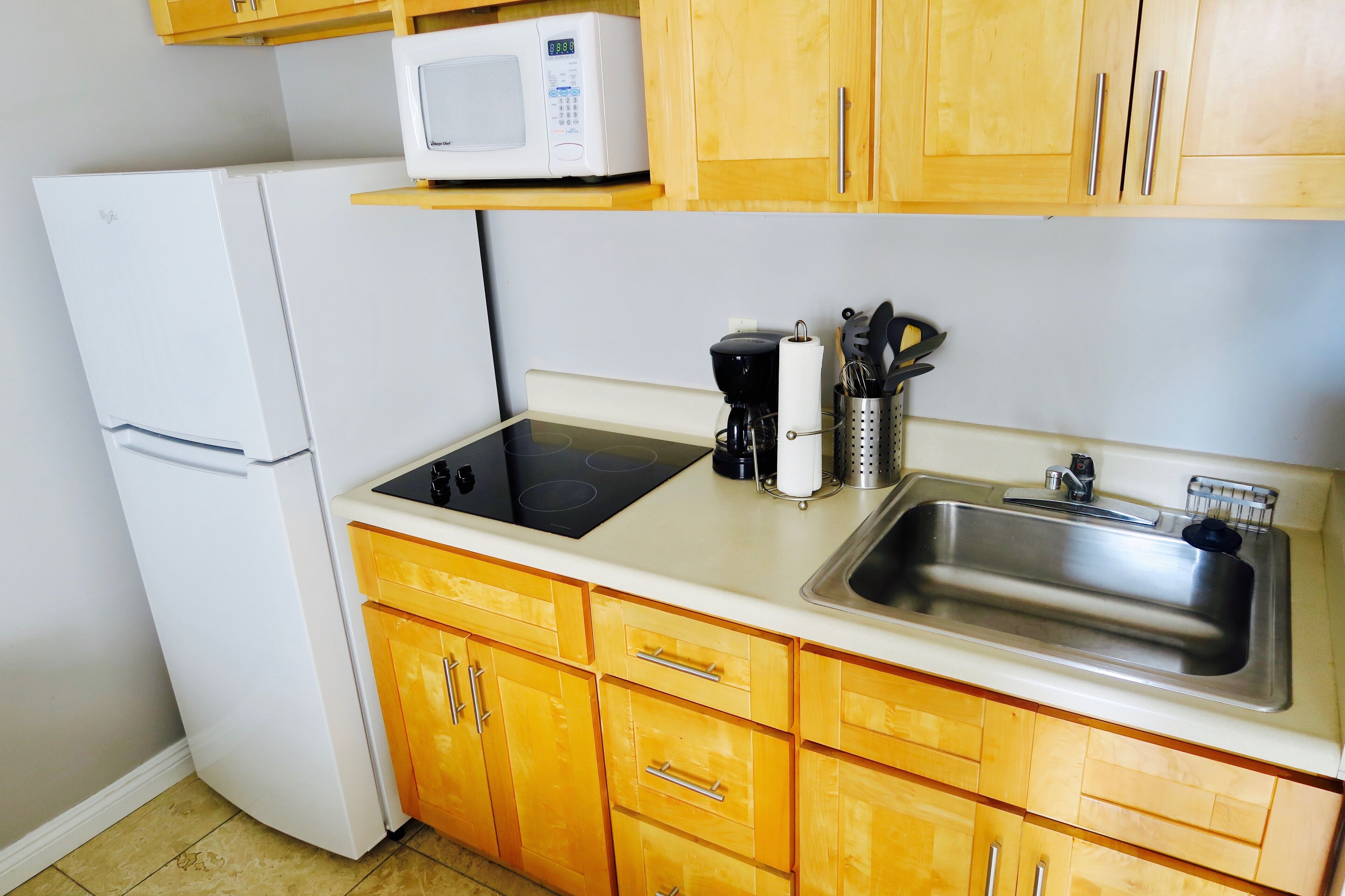 A compact kitchen with a white refrigerator, a microwave on a shelf, a black electric stove, a stainless steel sink, and wooden cabinets.