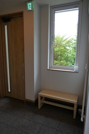 A room with a wooden bench, a window with a view of greenery, a door, and a wall-mounted emergency exit sign. - Bliss & Bless Furano (Furano)