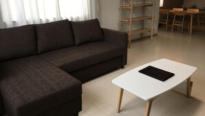 A modern living room with a dark brown sectional sofa, a white coffee table, and a wooden shelving unit. - Bliss & Bless Furano (Furano)