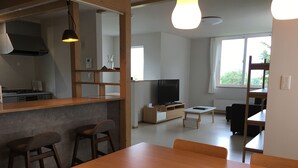 A modern kitchen with a built-in stove, a dining area with wooden chairs and a table, a TV on a wooden stand, and a large window with a view of greenery. - Bliss & Bless Furano (Furano)