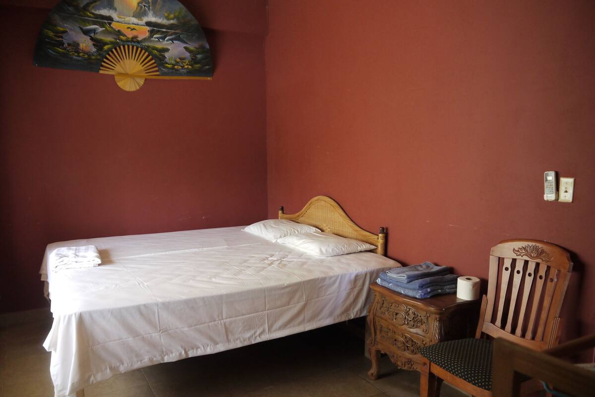 A single bed with a wooden headboard, a small bedside table with a lamp, a chair, and a wall-mounted painting.