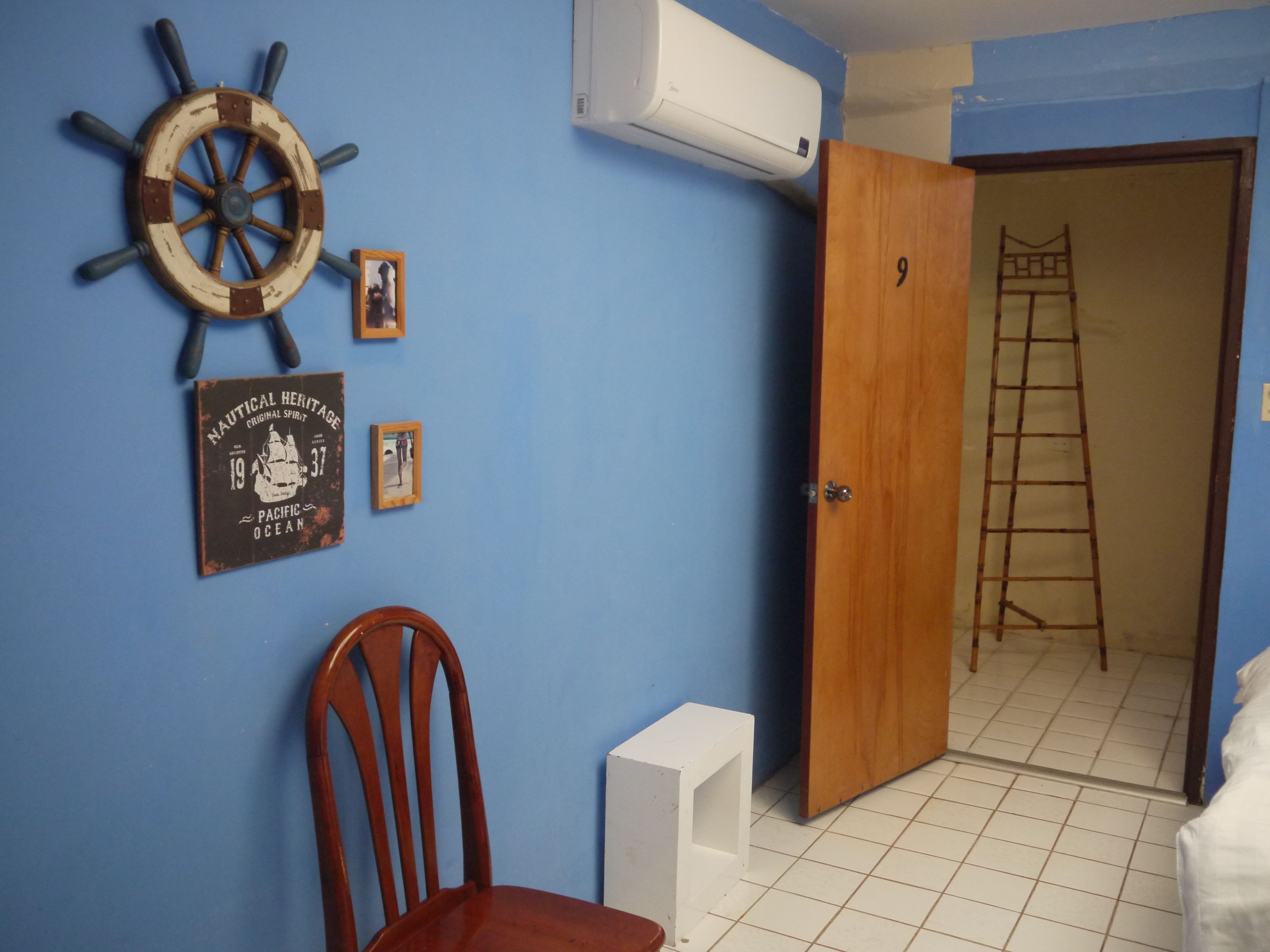 A room with a wooden door, a white air conditioner, a wooden chair, a small white table, and a wall with nautical-themed decorations.