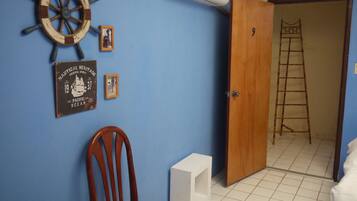 A room with a wooden door, a white air conditioner, a wooden chair, a small white table, and a wall with nautical-themed decorations.